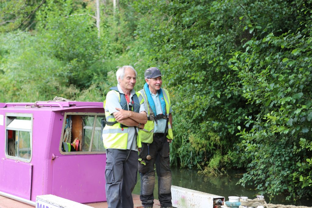 two male volunteers stood smiling on a canal boat while it travels down the canal.