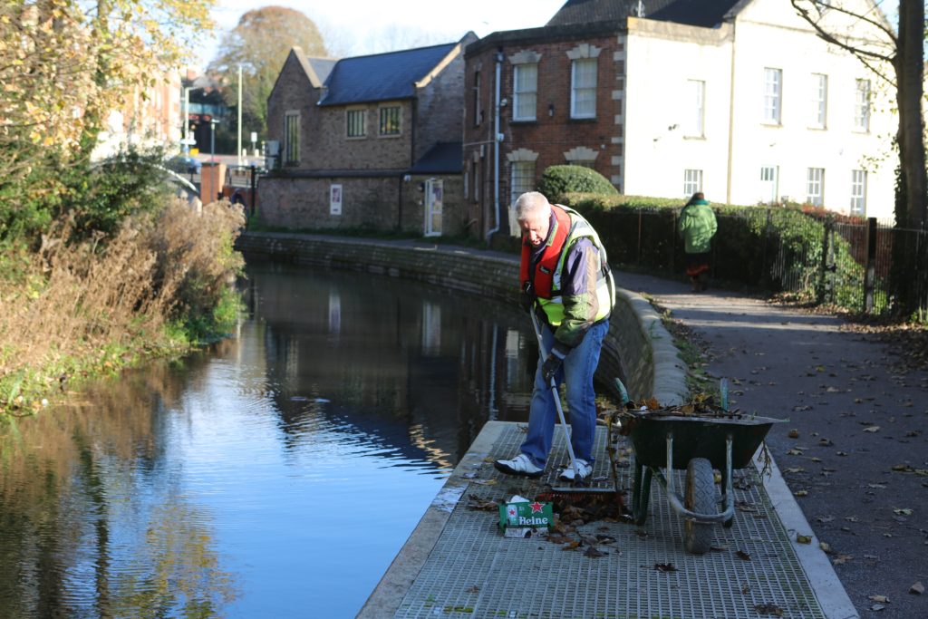Pictured is a male volunteer in life-jacket and high visability clothing. He is sweeping up litter next to the canal. There is a wheelbarrow next to his right.